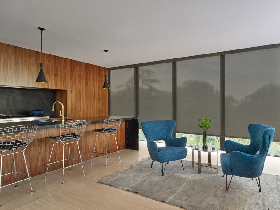 Designer Roller Shades covering floor to ceiling windows in a modern kitchen with dark hardwood. There is a sitting area with blue statement chairs and a small table in between them.